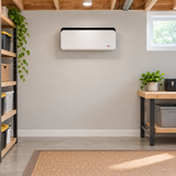 A white wall-mounted dehumidifier with a black top trim, installed on a light gray wall in a home office or garage setting with wooden ceiling beams, shelving, plants, and a workbench.