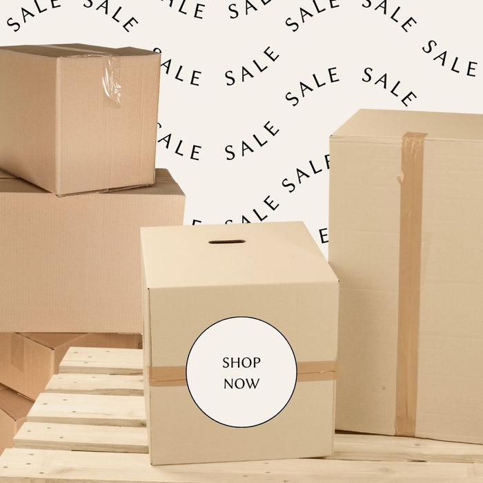 A stack of plain cardboard boxes on a light wood surface, with a central box featuring a round white label that reads “SHOP NOW.” The background is white with a repeating “SALE” text pattern.