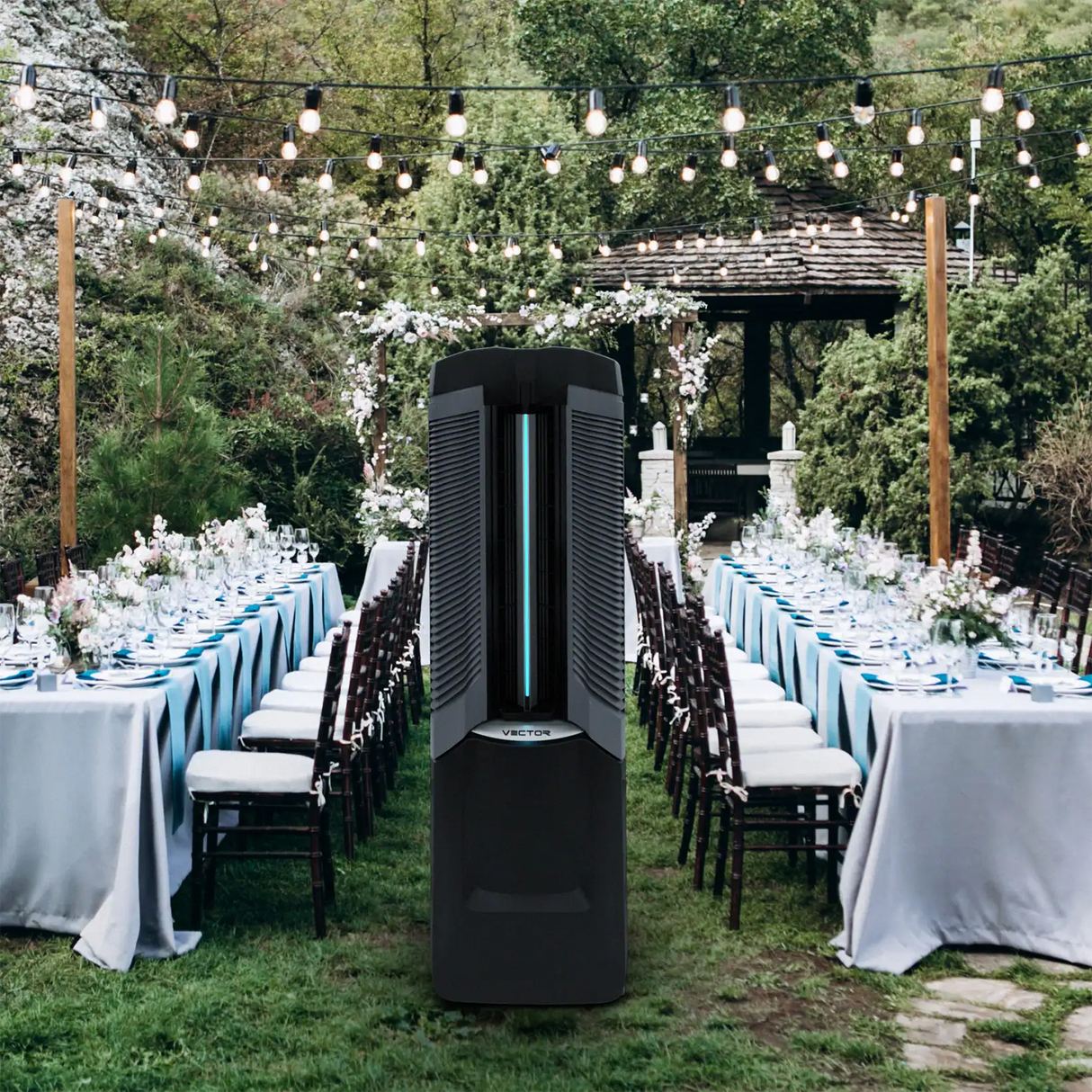 A tall black freestanding Vector 9,000 CFM Climate Control System  stands on grass in an outdoor garden event setting. Long tables with white and light blue linens, chairs, and string lights are arranged in the background.