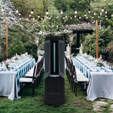 A tall black freestanding Vector 9,000 CFM Climate Control System  stands on grass in an outdoor garden event setting. Long tables with white and light blue linens, chairs, and string lights are arranged in the background.