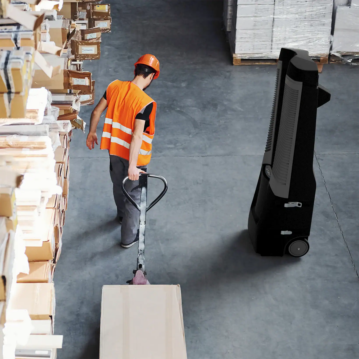 Vector 9,000 CFM Climate Control System cooling a warehouse. A warehouse worker in an orange safety vest and hard hat pushes a pallet truck carrying a box, with stacked cardboard boxes on the left and a black wheeled cart on the right.